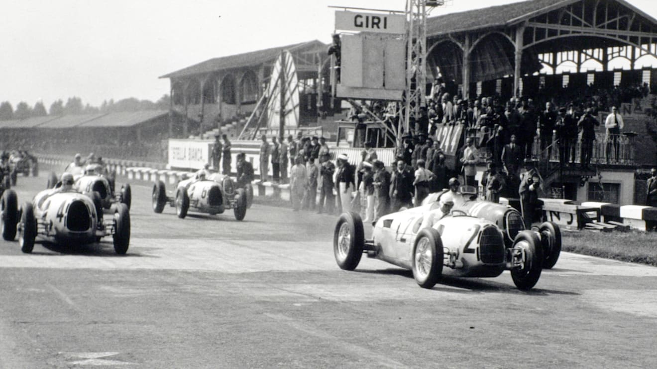 Hans Stuck Snr, in the #2 Auto Union Type C, leads the start of the 1936 Italian Grand Prix at Monza from Bernd Rosemeyer, #4 Auto Union, Tazio Nuvolari, #18 Alfa Romeo, Achille Varzi, #6 Auto Union, Ernst von Delius, #8 Auto Union, and Giuseppe Farina, #22 Alfa Romeo