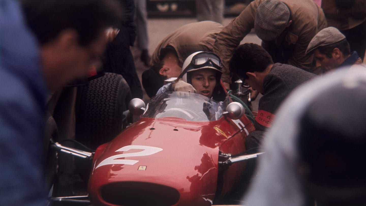 John Surtees in a Ferrari. Viewed through a crowd of people, with mechanics working on his car.